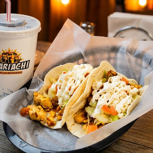 Two chicken tacos with lettuce, tomatoes, and crema, next to an El Mariachi drink cup.