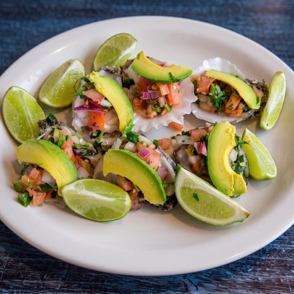 Oysters topped with fresh salsa and avocado slices, garnished with lime wedges on a white plate.