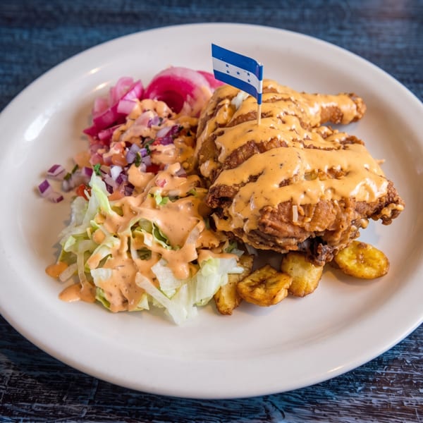A plate of Honduran fried chicken, salad, plantains, and pickled onions, topped with sauce and a flag.