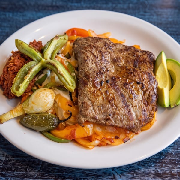 Grilled steak with sautéed peppers, onions, avocado, and refried beans on a white plate.